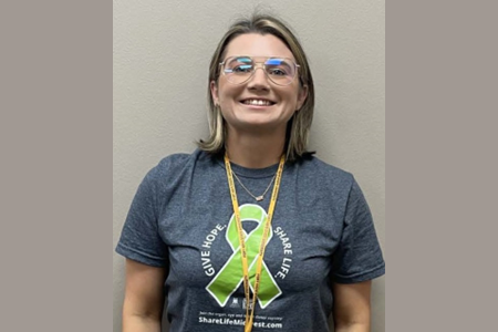 A woman wearing glasses, smiling for a picture, wearing a T-shirt with a green ribbon in support of organ donation.